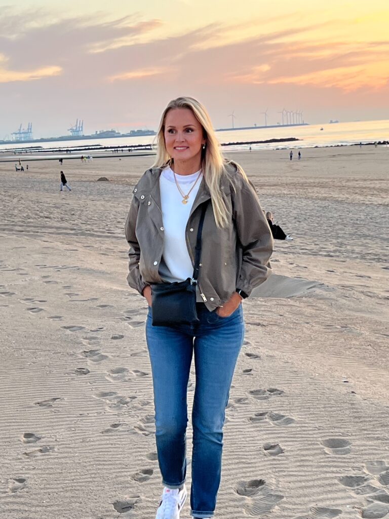 Maddy Willems wandelt op het strand bij zonsondergang, met een ontspannen glimlach en een kleurrige lucht op de achtergrond.
