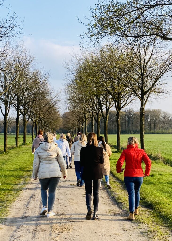 Groep vrouwen wandelt in een rij bomen op een zandweg in het groen, onder een blauwe hemel in voorjaarslicht.